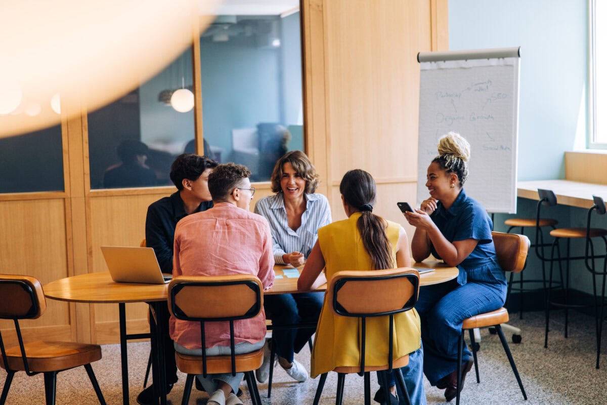 A diverse group of individuals gathered around a table, engaging in an active discussion with notes on a flipchart in a well-lit, modern workspace environment.